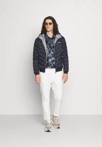 Navy Pufferjacke mit grauem Innenfutter, Batikshirt, weißen Hosen und beigen Turnschuhen. Zubehör sind gelbe Sonnenbrillen.