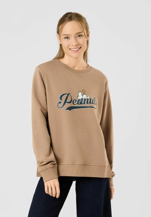 Jeune femme souriante portant un sweat-shirt beige avec un logo et un personnage des Peanuts, associé à un pantalon foncé sur un fond gris clair uni.