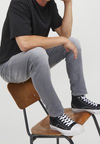 Vaqueros de mezclilla gris de corte ajustado, combinados con zapatillas de lona negras de caña alta con cordones blancos, sentados en una silla de madera.