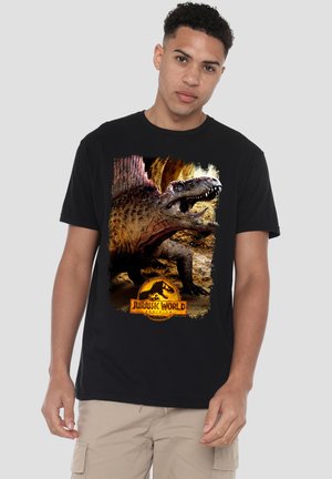 Junger Mann trägt ein schwarzes Jurassic World T-Shirt mit einem brüllenden Dinosaurier und Logo, steht vor einem einfarbigen hellgrauen Hintergrund.