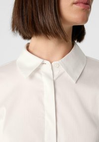 Ein weißes Hemd mit Kragen aus glattem, strukturiertem Stoff, das über eine verdeckte Knopfleiste und ein klassisches Kragendesign verfügt und einen sauberen Look kreiert.