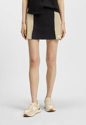Black and beige sports skirt with side panels, accented with white piping and a logo on the front. Worn with beige sneakers.