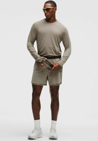 Sportos öltözet, amely hosszú ujjú, világosbarna pólót, illő sortot és fehér sportcipőt tartalmaz; egy logós vízpalackot tart a kezében.