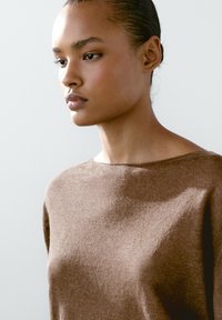 Hnědý pletený svetr s širokým výstřihem, měkkou texturou a volným střihem. Detail obličeje modelky s hladkou pletí a staženými vlasy.