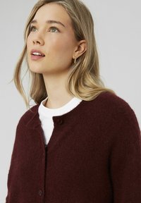 Cardigan en tricot bordeaux avec un col rond, une fermeture à un bouton et une texture douce, superposé sur une chemise blanche.