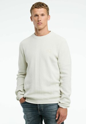 Lichtbeige gebreide sweatshirt met ribbeltextuur, ronde halslijn en een klein logo-accent. Gepaard met blauwe spijkerbroeken.