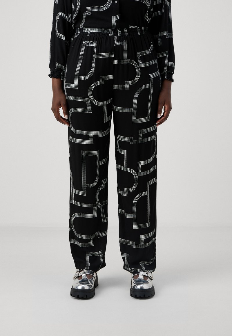 Pantalones negros con un patrón que presenta formas geométricas blancas en contraste, con una cintura elástica, bolsillos laterales y un corte relajado. Combinados con zapatos plateados brillantes.