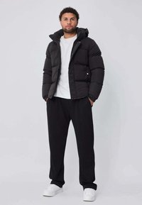 Doudoune noire avec capuche, présentant des sections matelassées. Associée à un t-shirt blanc et un pantalon noir, portée avec des baskets blanches.