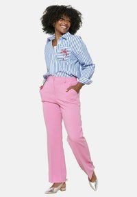 Črtasta modro-bela majica z žepom v obliki palme, v kombinaciji z roza hlačami in srebrnimi nizkotenimi čevlji. Kudiraste lase, roke v žepih.