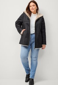 Zwarte ritsjack met een faux fur gevoerde capuchon, met een losse pasvorm en zijzakken, gedragen over een witte coltrui en blauwe jeans.