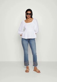 Blusa branca texturizada com decote quadrado e mangas bufantes, combinada com jeans azuis com detalhes bordados na bainha e sandálias.