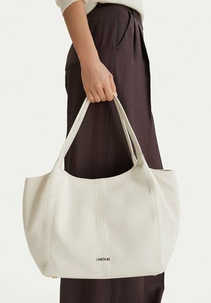 Hand hält große cremeweiße Ledertasche mit Doppelgriffen und LASOCKI-Logo, getragen mit weißem Pullover und dunkler Hose.