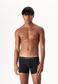 Black cotton boxer briefs with a logo waistband, featuring a fitted shape and smooth texture, model standing against a plain white background.
