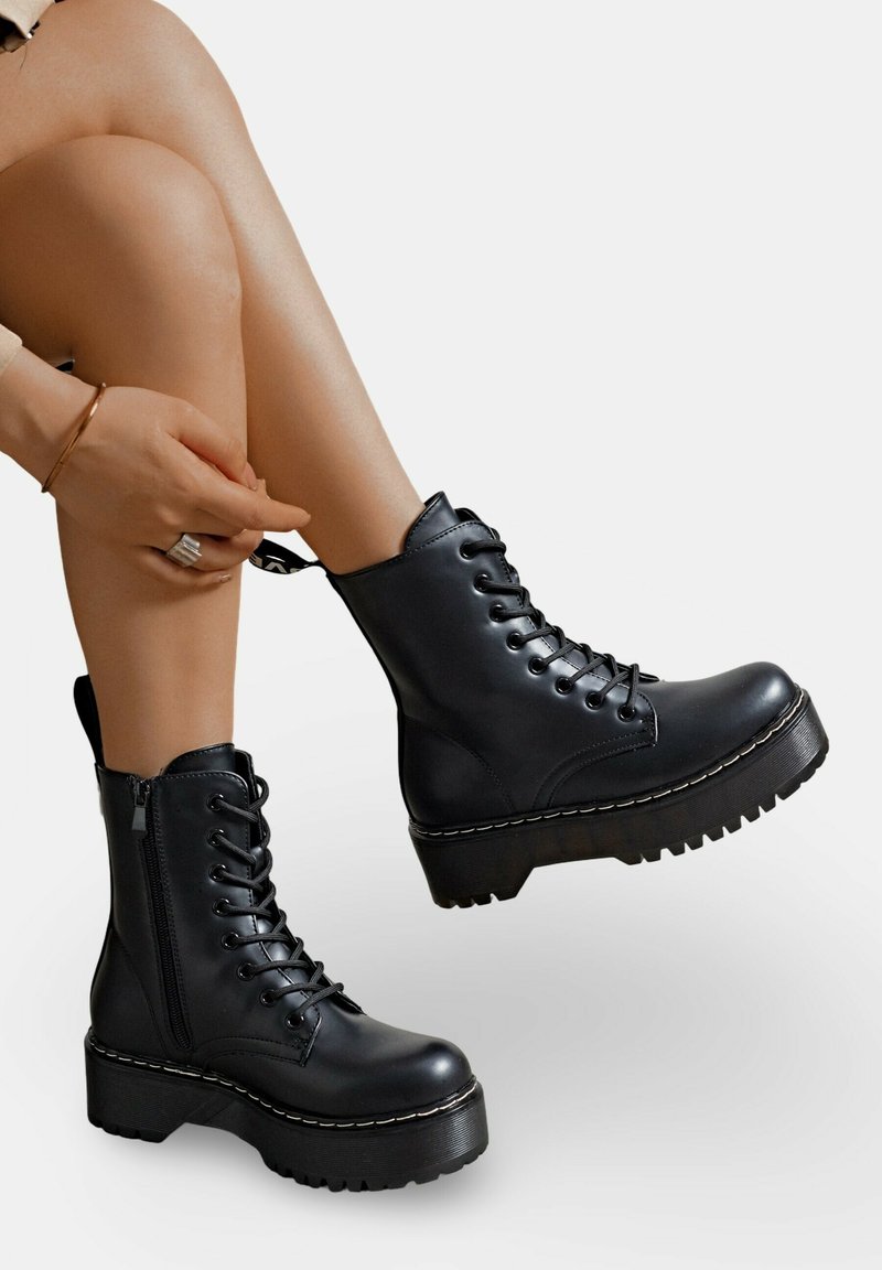 Bottes noires en cuir à la cheville avec un laçage à l'avant, une fermeture éclair sur le côté, une semelle en caoutchouc épaisse et un détail de couture blanche contrastante.