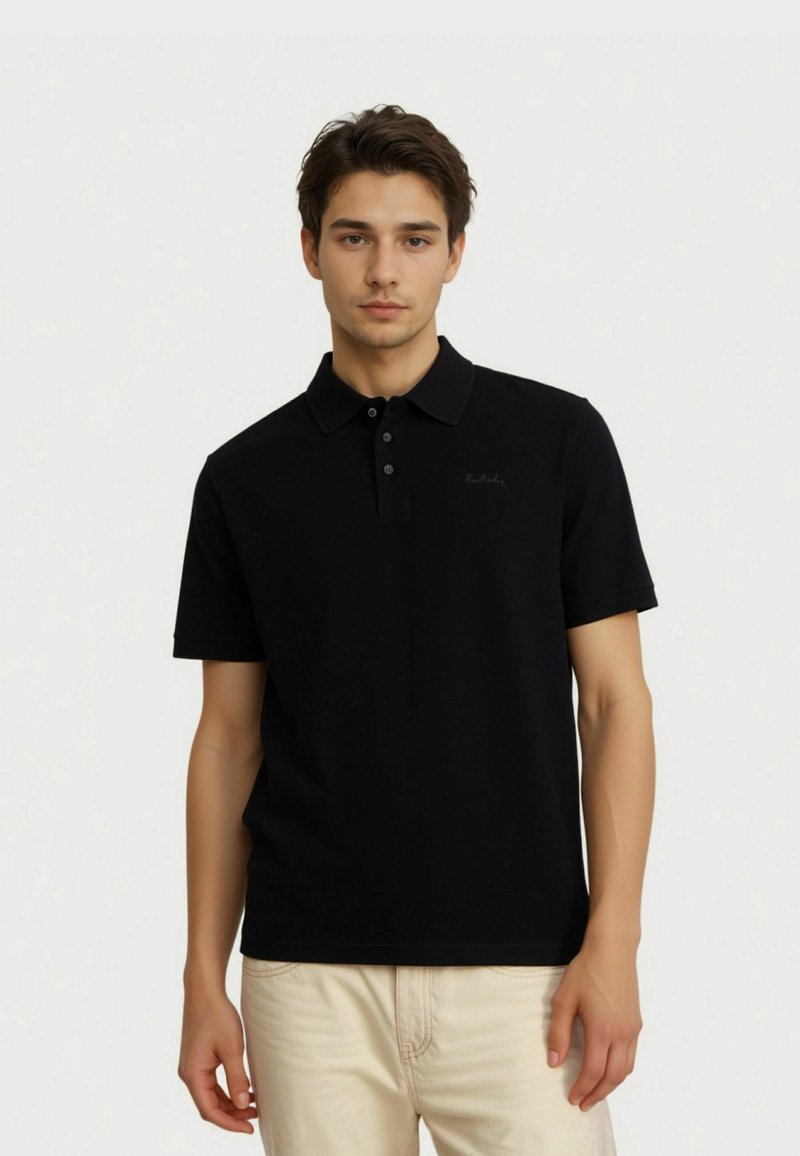 Musta poolpaita lyhyillä hihoilla, jossa on kolme nappia, pehmeä tekstuuri ja hienovarainen brändin brodeeraus rinnassa, yhdistettynä vaaleisiin housuihin.