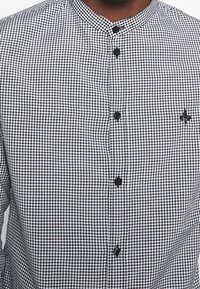 Camisa a cuadros en blanco y negro con cuello de banda, botones negros y un pequeño detalle bordado de flor de lis en el pecho.