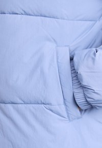 Veste matelassée bleu clair avec une texture lisse, dotée d'une poche latérale et de poignets froncés pour un ajustement snug. Détails de design minimalistes.