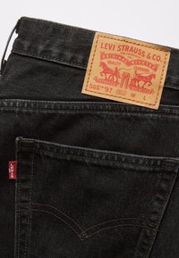 Jeans en denim noir avec l'étiquette rouge Levi's sur la poche et une étiquette en cuir marron de Levi Strauss & Co. montrant des chevaux et le texte "565 '97 Coupe droite ample."
