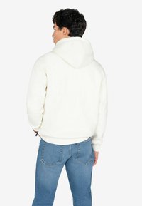 Sudadera blanca con capucha y cordón, vista desde la parte trasera, combinada con jeans de color azul claro, con un ajuste relajado y puños acanalados.