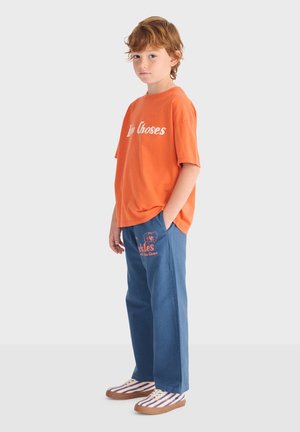 Jeune personne debout, portant un t-shirt orange "My Choses", un pantalon bleu avec des inscriptions imprimées, et des baskets rayées, les mains dans les poches.