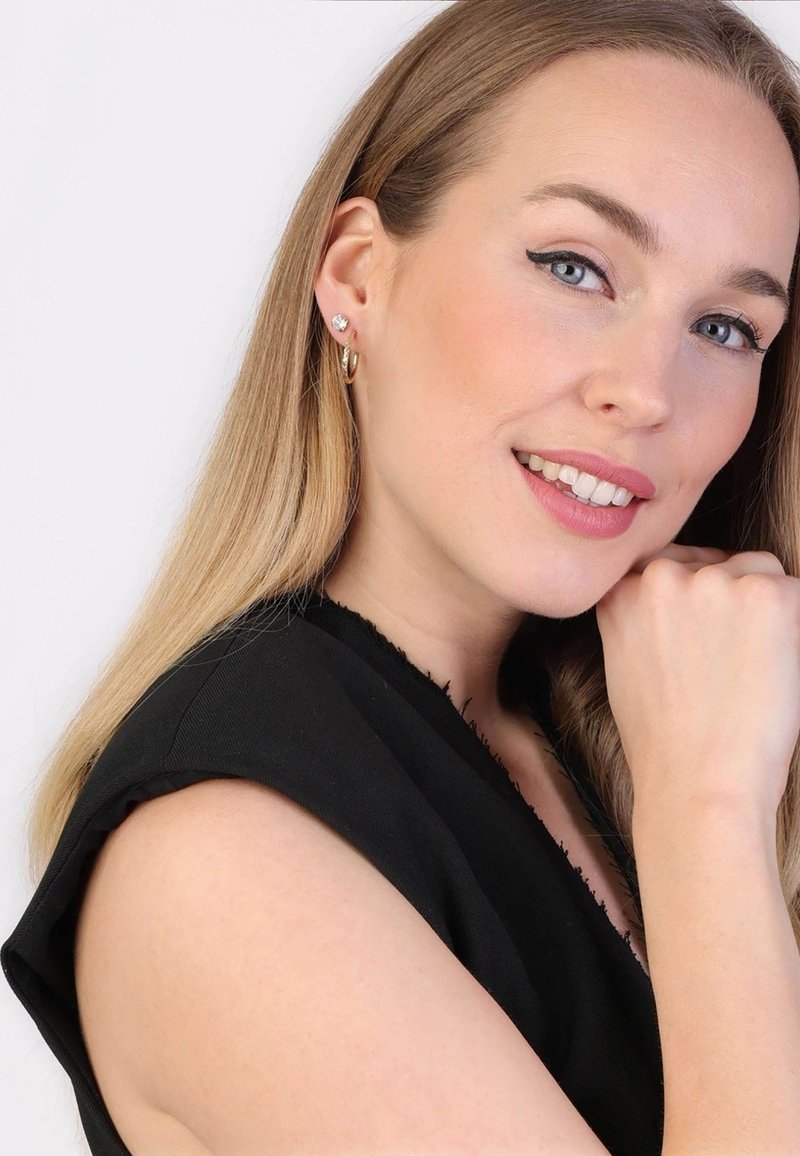 Gouden hoepeloorbellen en een studsieraad met een heldere steen, gedragen door een vrouw met lang, steil blond haar en een lichte huid, gekleed in een zwarte top.
