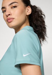 Světlá tyrkysová polo košile s krátkými rukávy, s bílým logem Nike na levém rukávu. Hladký materiál a klasický design límce.