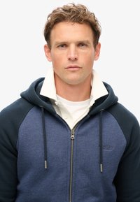 Sudadera con cremallera de color azul marino y un interior gris claro, con un cuello blanco debajo y detalles de cordones. Logo "SuperDry" en la parte frontal.
