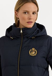 Tamsiai mėlyna pūkinė striukė su kapišonu, su auksinės spalvos užtrauktuku ir logotipo emblema. Lygaus audinio, su siuvinėtomis dalimis šilumai užtikrinti.