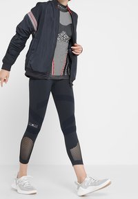 Meriini pommitusjakku punaste ja valgete triipudega, hall mustriga ilma varrukateta topp, mustad retuusid võrgusilmaga aktsentidega ja hallid spordijalatsid.