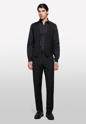 Completo nero con gilet su misura dotato di bottoni, camicia a maniche lunghe e pantaloni affusolati, il tutto realizzato in un tessuto morbido, completato da scarpe lucide.