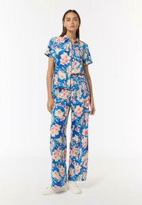 Outfit a fiori nei toni del blu, rosa e crema. Camicia a maniche corte con bottoni abbinata a pantaloni lunghi a gamba larga. Sneakers bianche.