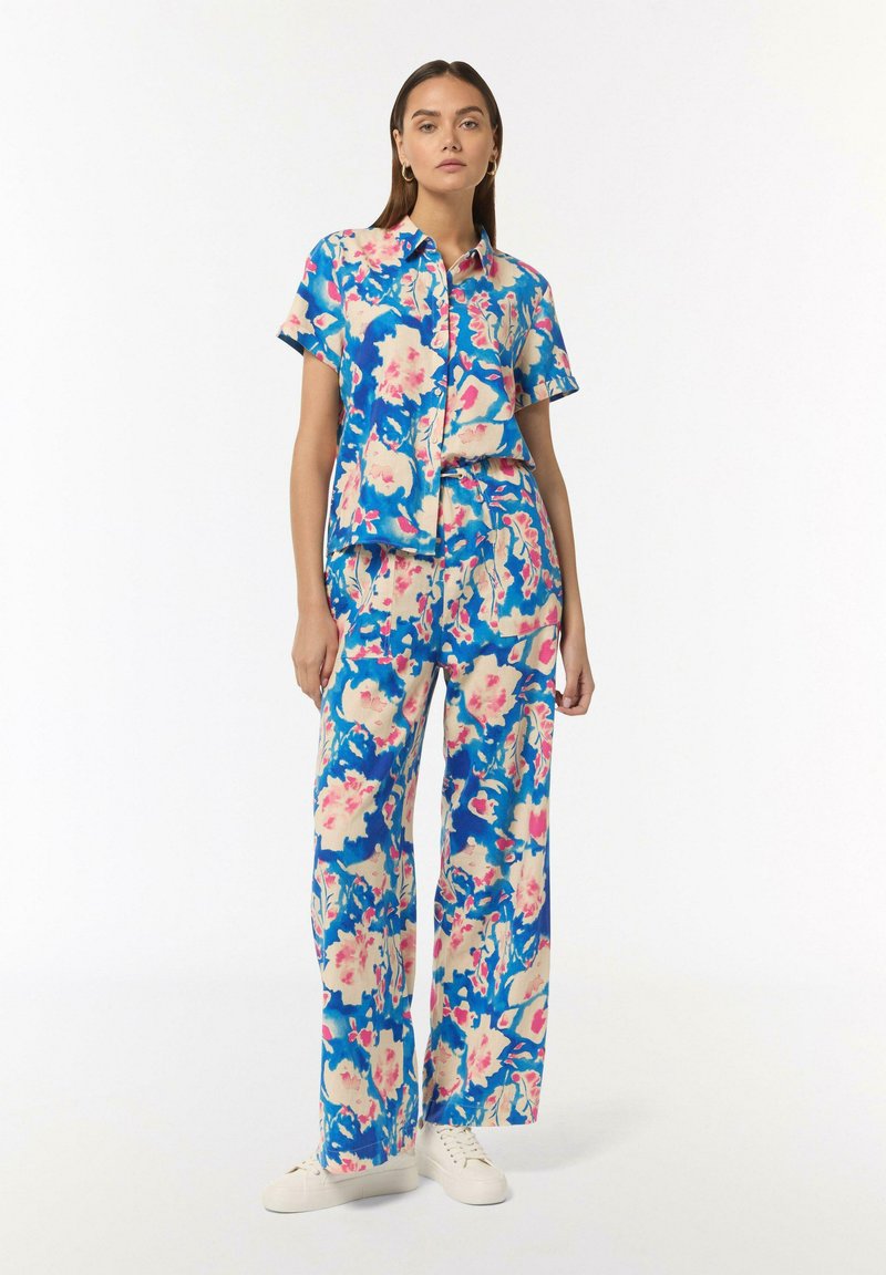 Outfit a fiori nei toni del blu, rosa e crema. Camicia a maniche corte con bottoni abbinata a pantaloni lunghi a gamba larga. Sneakers bianche.