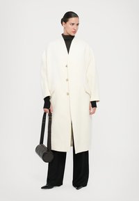 Long white coat with a relaxed fit, three buttons, and side pockets, paired with black pants and a brown crossbody bag.