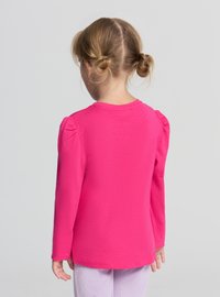 Różowa bawełniana koszula z długim rękawem z marszczonymi bufkami oraz okrągłym dekoltem, noszona przez dziecko zwrócone tyłem, w połączeniu z jasnymi spodniami w paski.