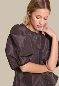 Mørkebrunt, shiny stoffetopp med puffermer og rund halslinning. Designet har en jevn tekstur og subtile samlinger ved ermene.