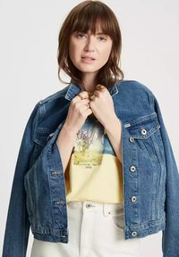 Niebieska dżinsowa kurtka o lekko skróconym kroju, z zapięciem na guziki i bocznymi kieszeniami, noszona na żółtej koszulce z grafiką i białych dżinsach.