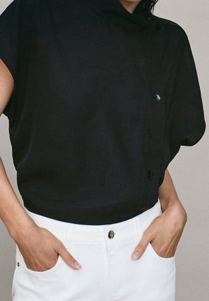 Personne portant un haut noir ample avec des boutons et un pantalon blanc, les mains dans les poches, sur un fond uni.