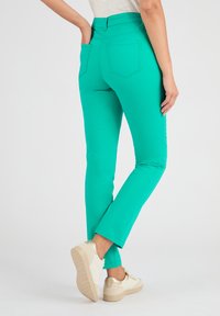 Pantaloni elasticizzati verdi con una vestibilità slim, dotati di tasche posteriori e una texture liscia. Abbinati a scarpe bianche con un accento dorato.