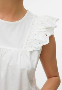 Blouse blanche avec un col rond, dotée d'épaules en dentelle froncées et d'un corsage plissé. Tissu en coton lisse avec des détails élaborés.