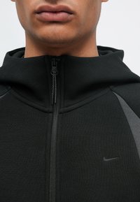 Černá mikina na zip s vysokým límcem, s šedými akcentními panely na ramenou a vyraženým logem Nike na hrudi. Měkká textura.