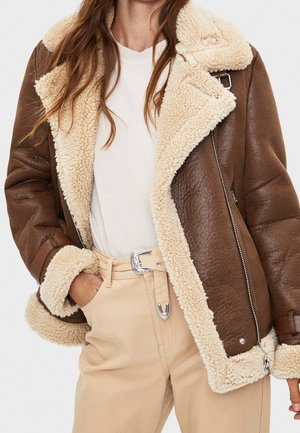 Vrouw met een bruine shearling jas met roomkleurige fleece voering, witte blouse en beige broek met een zilverkleurige gesp, hand in de zak.