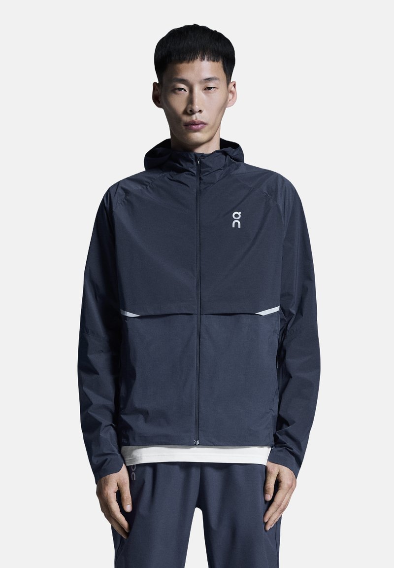 Veste coupe-vent bleu marine foncé avec capuche, dotée d'une fermeture éclair frontale, d'accents réfléchissants et d'un logo sur la poitrine. Tissu lisse et léger.
