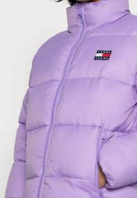 Osoba w jasnofioletowej, pikowanej puchowej kurtce z wysokim kołnierzem i logo Tommy Jeans na piersi, z rękami w kieszeniach.