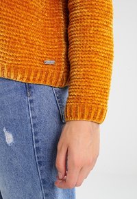 Oranger, strukturierter Samtpullover mit gerippten Bündchen, getragen über abgenutztem blauen Denim, mit einem kleinen Metall-Logoanhänger.