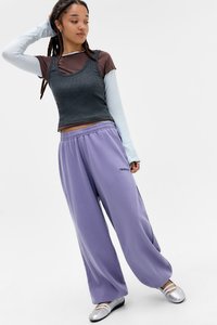 Top cinza sem mangas sobre uma camisa de manga longa marrom, combinado com calças de moletom largas roxas e sapatilhas prateadas.