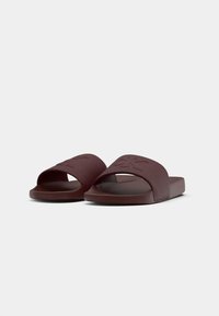Sandales à glissière bordeaux avec une sangle texturée et des semelles plates. Le design présente un logo en relief sur la sangle, soulignant un style minimaliste.