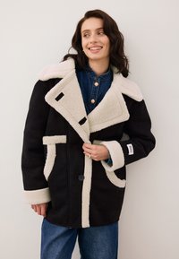Czarny płaszcz z wełny z domieszką z kremowym kołnierzem i mankietami z syntetycznego owcza, kontrastowymi wykończeniami, przesadnymi klapami i pojedynczym zapięciem na guzik.