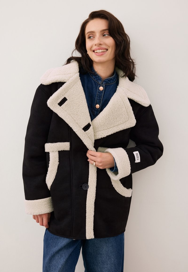 Czarny płaszcz z wełny z domieszką z kremowym kołnierzem i mankietami z syntetycznego owcza, kontrastowymi wykończeniami, przesadnymi klapami i pojedynczym zapięciem na guzik.