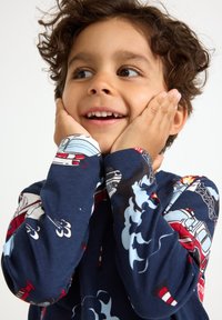 Ein curlyhaariges Kind lächelt mit den Händen auf den Wangen, trägt ein dunkelblaues Langarmshirt mit Feuerwehrwagen und Rauchmustern.