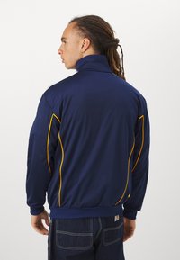 Veste de sport bleu marine avec col montant, accents jaunes le long des coutures, poignets et ourlet côtelés, fabriquée en tissu lisse et léger.